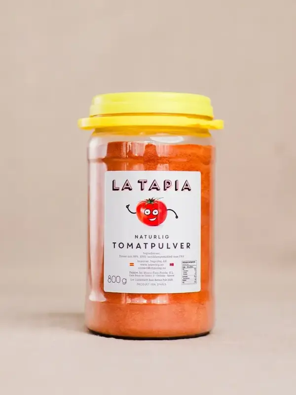 Beholder med La Tapia tomatpulver, plassert ved siden av frisk tomat på kjøkkenbenken.